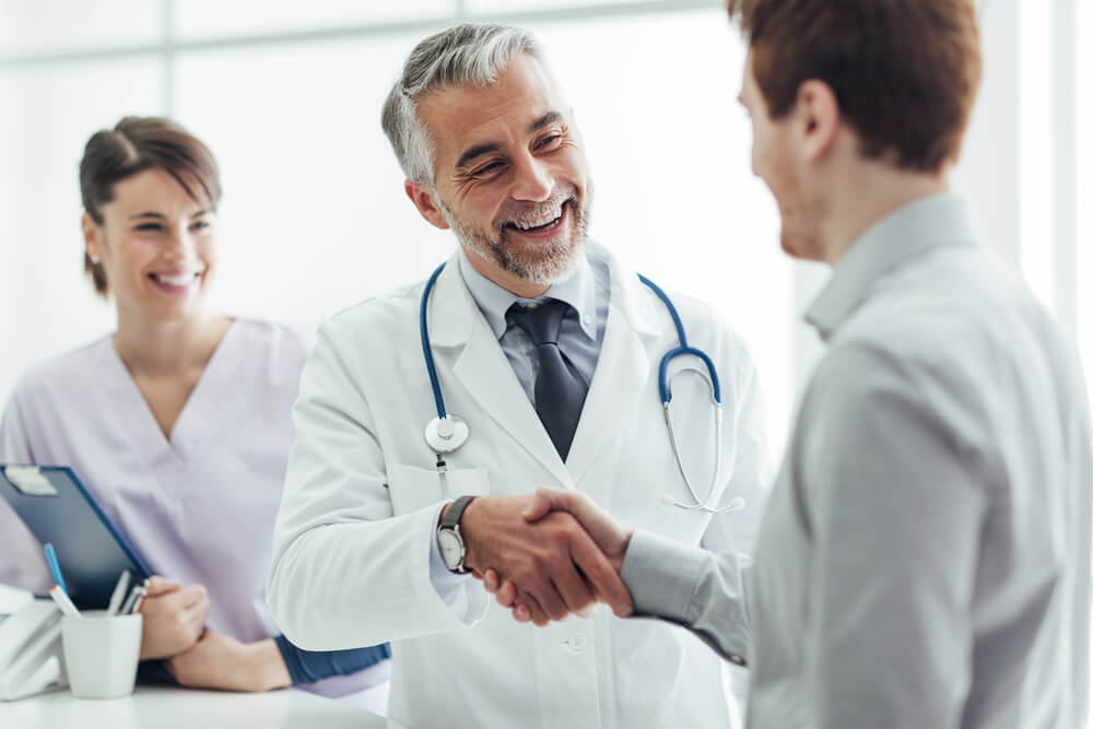 A smiling doctor wearing a white lab coat, tie, and stethoscope shakes the hand of a man in a gray shirt. In the background is a woman in lavender scrubs holding a clipboard.