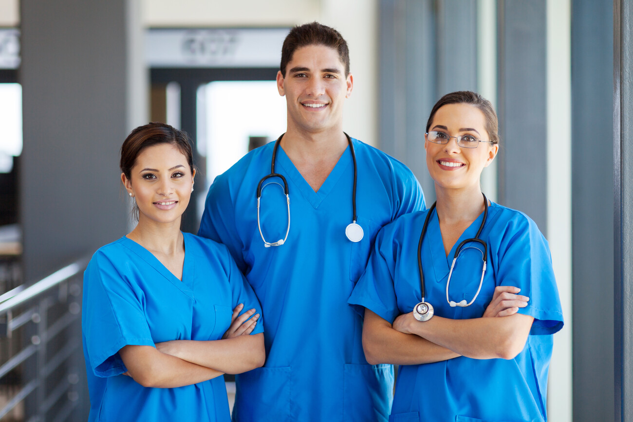 Two women and one man wearing blue scrubs stand together, smiling. Two of them are wearing stethoscopes.