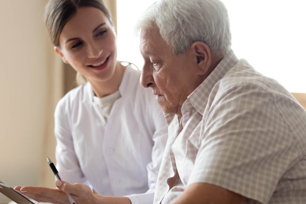 A woman in a white coat sits next to an older man and smiles at him. She holds a clipboard and a pen.