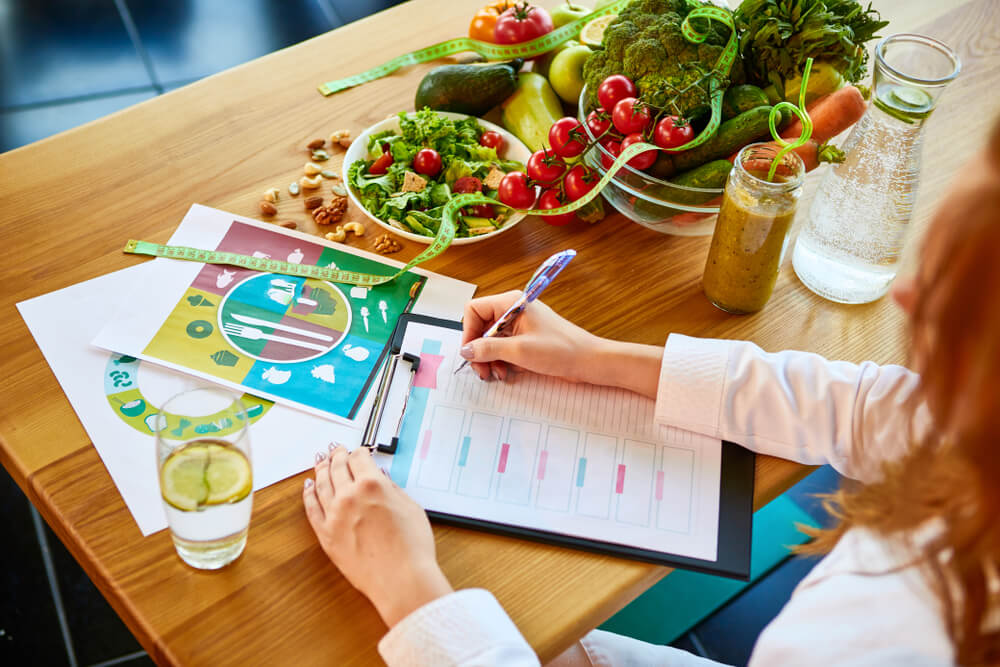 A woman in a white lab coat sits at a table with a bowl of vegetables, salad, nuts, and glasses of juice and water. She appears to be writing a meal schedule.